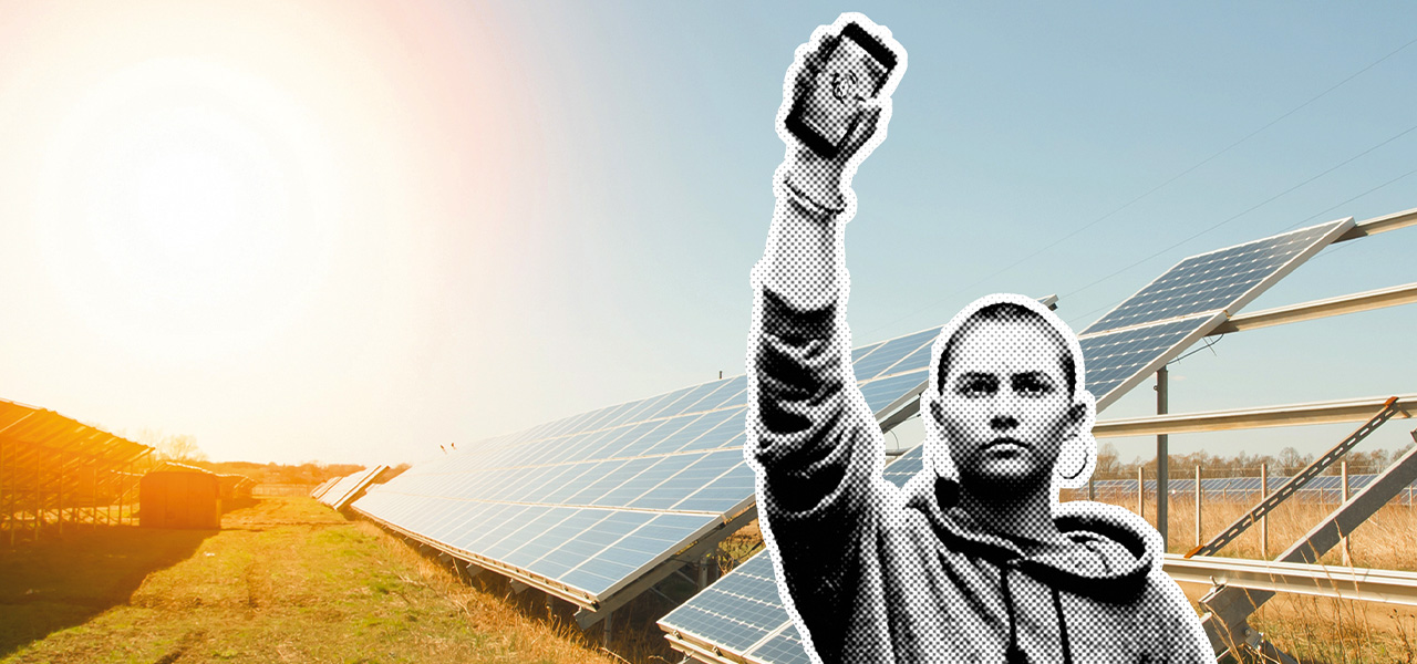 Eine Frau steht vor einer Photovoltaik-Freiflächenanlage. Sie hält ein Smartphone in die Höhe.